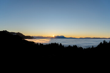 sunset over the summits of high mounatins above a sea of fog towards clear blue and orange sky in late autumn