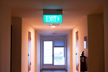 Green Illuminated Exit Sign Above Corridor Leading To Large Window With Natural Light
