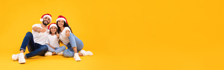 A cheerful family of three shares joyful moments during the Christmas season, wearing Santa hats and sitting on the floor with bright smiles against a vibrant yellow backdrop.