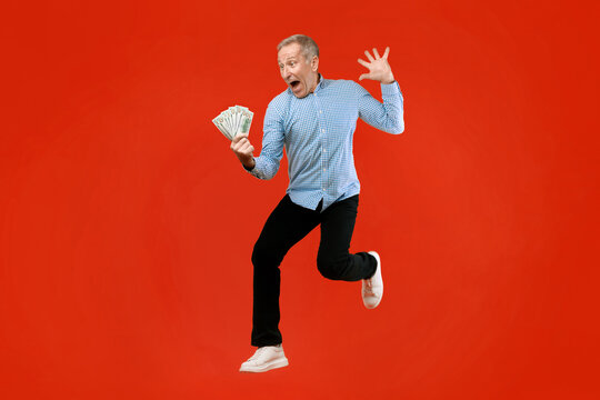 An older man jumps in the air with joy while holding a bundle of cash. He wears a blue checkered shirt and black pants with white shoes.