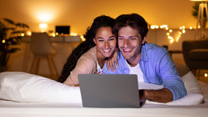 Happy spouses are lying in bed, watching a comedy movie on a laptop. They are enjoying each other's...