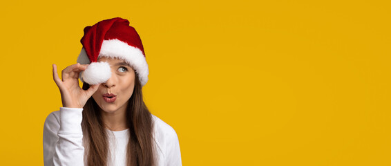 A woman with long hair is wearing a red Santa hat and holding a white Christmas ornament up to her eye. She has a playful expression against a cheerful yellow background, evoking holiday joy.