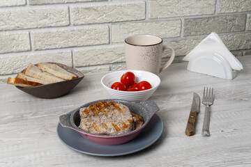 Breakfast table setting with bread, tomatoes, pork chops and a drink in a simple kitchen environment