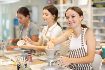 Teen girl student sculpting bowl of gray clay, smoothing surface of clay product with fingers and cutter. Handmade manufacturing from clay, hobby and leisure activity
