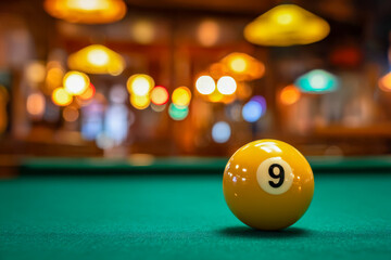 Yellow number nine billiard ball resting on green felt pool table in a cozy bar with warm glowing hanging lights creating a relaxed atmosphere