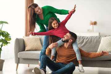 Positive Arab parents and their daughter share joyful moments in their cozy living room. The dad plays with their daughter while the mom smiles, creating a loving and fun family atmosphere.
