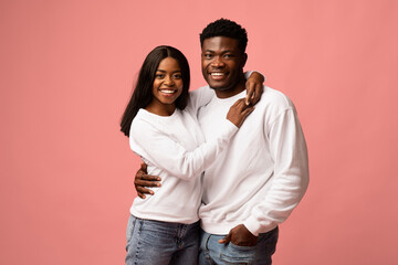Obraz premium A cheerful young black couple wearing white sweatshirts and jeans is embracing on a pink backdrop. They are smiling at the camera and enjoying their time together, showcasing their love.