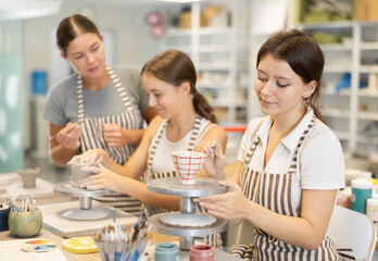 During workshop at master class, female pupil paints clay vessel, applying pigment to plate with brush. In background, mentor teaches student, helps to color bowl.