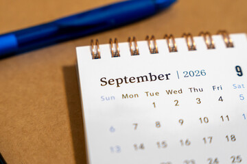 Detailed view of a desk calendar page displaying September 2026, accompanied by a blue pen, against a brown surface, suitable for planning, organization, and scheduling contexts.