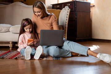 A mother and her young daughter are sitting on a rug in their living room. The mother is using a laptop, while the daughter is engaged with a smartphone. They are enjoying quality time together.