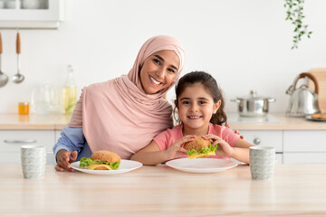 A mother in a light pink hijab and her daughter are happily posing with homemade burgers on their plates. The cozy kitchen is filled with cooking tools and a warm atmosphere.