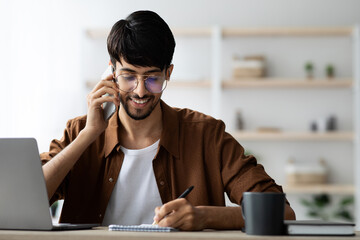 A young man sits at a desk with a laptop, smiling as he talks on the phone. He is writing notes in a notebook and enjoying a beverage, surrounded by a tidy workspace.