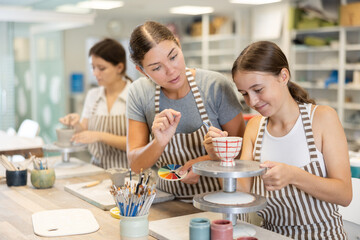 Young female teacher explains to teenage girl student how to paint ceramic cup in workshop