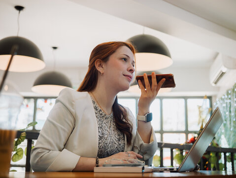 Focused woman dictating a voice message on her phone next to a laptop at a cafe table