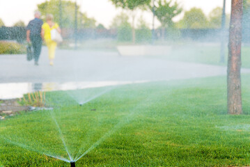 Sprinkler spraying water against park with man and woman walking