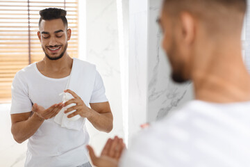 Fototapeta premium A man smiles as he applies moisturizer to his hands while standing in front of a mirror in a bright and modern bathroom. The atmosphere feels calm and refreshing.