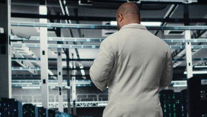 Data center system administrator checking infrastructure diagnostics using tablet screen. IT specialist walking in high tech server room, examining data charts during system audit, camera B