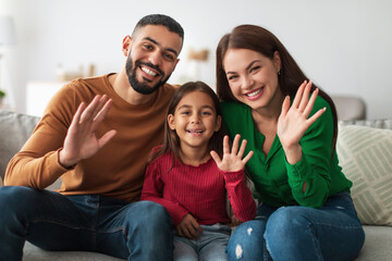 Fototapeta premium A cheerful family of three sits on a couch at home, waving and smiling at the camera.