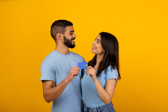 Two loving arab spouses embrace while holding a credit card. They share a moment of joy and connection, recommending a banking service against a bright yellow backdrop. - Powered by Adobe