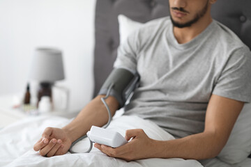 Fototapeta premium An Arab man sits on his bed, using an upper arm monitor to check his blood pressure. He appears to feel unwell as he monitors his arterial tension in a quiet bedroom.