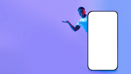 A young man stands beside a large blank screen, smiling and gesturing enthusiastically. The background features a vibrant purple hue, creating a lively atmosphere suited for presentations.