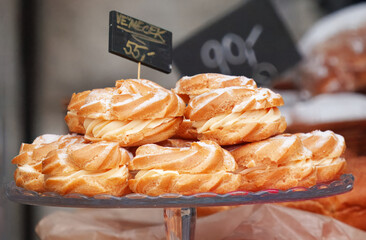 Golden swirled Czech cream pastries piled on glass cake stand at a lively farmers market in Prague. Flaky baked shells, silky egg yolk cream and dusting of sugar invite indulgence and festive delight.