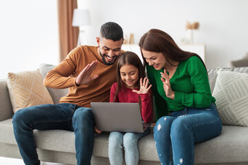 A joyful Arab family is engaged in a video call with their relatives, smiling and waving their hands. They sit comfortably on a sofa in a light and cozy living room, sharing happy moments together.