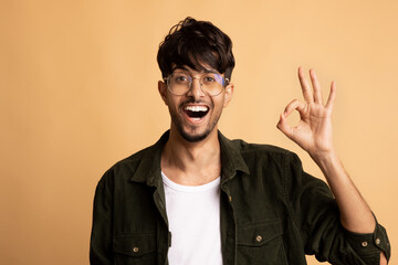 Naklejka premium A young man stands in front of a beige background, smiling widely and making an okay sign with his hand. He wears casual clothing and stylish glasses, highlighting a cheerful mood.