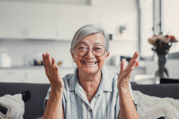 Senior woman sits on a sofa at home and smiles during an online video call. She enjoys connecting...