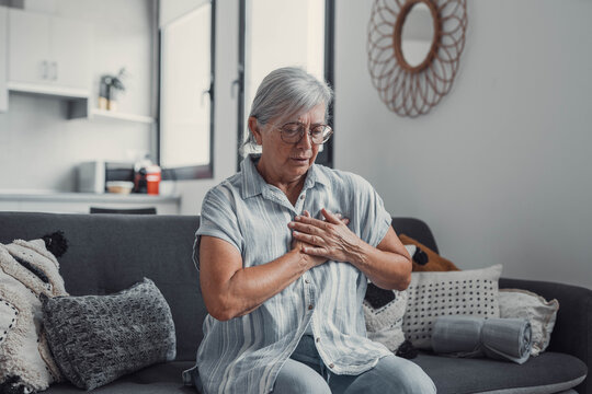 Elderly woman sits on a sofa at home holding her chest in discomfort. She appears worried and fatigued, showing signs of heart pain and anxiety during a stressful moment.