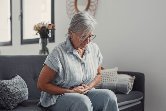 Elderly woman sits on a sofa at home holding her stomach, experiencing abdominal pain and discomfort. She looks tired and stressed while managing digestive issues during her daily life.