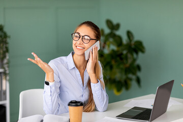 In a modern office, a happy businesswoman is talking on her cellphone and working on a laptop. She looks focused and engaged while chatting, with a coffee cup nearby.