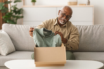 An elderly man is smiling while unpacking a freshly delivered shirt from a cardboard box in his...