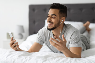 A happy young Arab man is lying on his stomach in bed, using a smartphone with earphones while making a video call at home. He is smiling and engaging in online conversation, showing delight.
