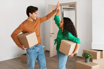 Young couple joyfully gives a high five while holding moving boxes in their new home. They are excited about their first home together, surrounded by unpacked belongings.