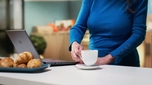 Senior adult in a calm kitchen setting scrolling apps on her laptop, enjoying peaceful retired life. Woman showing a gentle smile, simple moments that reflect relaxed domestic living. Camera B.