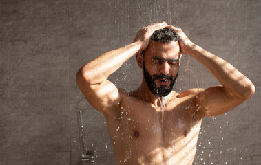 Young Middle Eastern man stands under a refreshing shower, eyes closed and hands in his hair. Water...