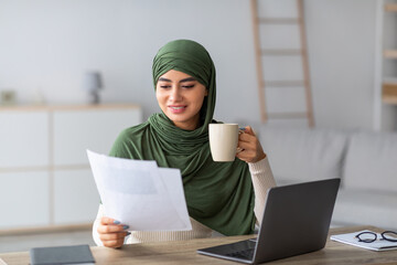 Confident young Arab lady in hijab sips hot coffee while reviewing business documents during an online work meeting at her home office. She manages her freelance career with focus and style.