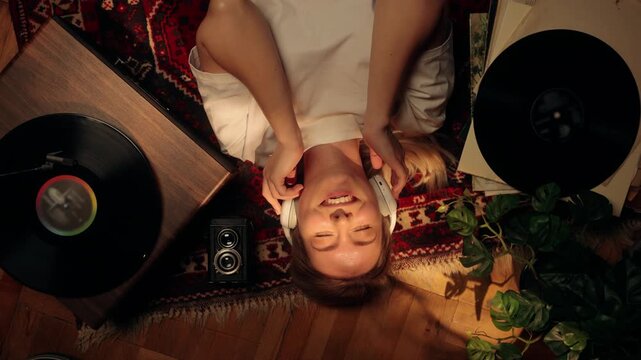 Young woman lying on the floor listening to music with headphones, smiling and relaxing at home. Cozy lifestyle scene with vinyl records, warm light, and calm atmosphere