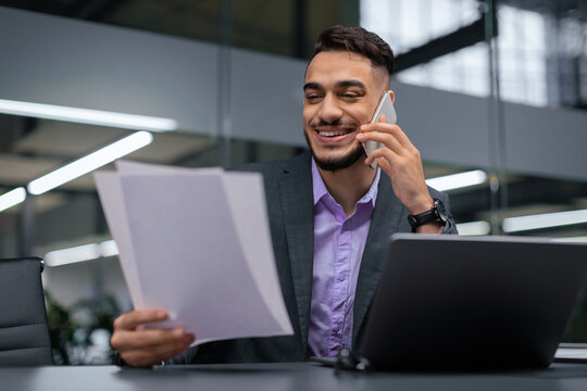 Man is talking on a phone while looking at a stack of documents in a modern office. He seems focused and happy while sitting at a desk with a laptop.