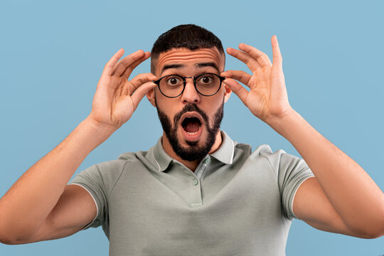 A man with a beard expresses surprise while adjusting his glasses. He holds them up with both hands, showcasing a moment of shock or excitement against a light blue backdrop.