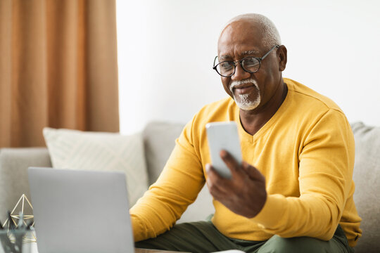 An older man is engaged in a digital activity, holding a smartphone while seated on a comfortable couch. A laptop rests in front of him in a warm, inviting living room. - Powered by Adobe