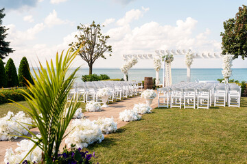 Beautiful outdoor wedding ceremony setup by the sea with white chairs, floral aisle decorations, and a pergola adorned with flowers, creating an elegant and romantic coastal atmosphere