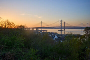 Evening settles across the Forth. The bridges gleam as water reflects warm light.