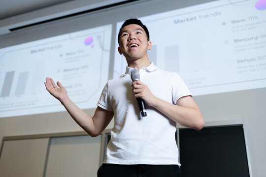An Asian male student holds a microphone and gestures while delivering a presentation in front of a projection screen. He appears confident and engaged while speaking to his class.