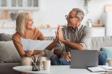 An older couple shares a joyful moment at home discussing plans. They sit on a cozy couch surrounded by papers and a laptop, showcasing their lively interaction and connection. © Prostock-studio