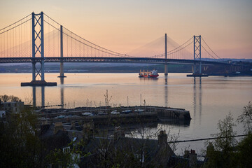 Obraz premium Warm sunset paints the Firth of Forth as the Forth Bridges rise above calm water. A small vessel sails past South Queensferry with Edinburgh on the horizon.