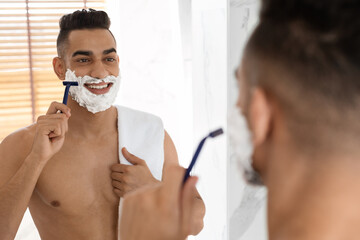 A young man with a towel around his body is smiling while shaving in front of a bathroom mirror. He has shaving cream on his face and is enjoying the grooming process.