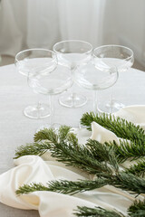 Minimalist holiday table setting with elegant coupe glasses, natural pine branches and soft white fabric, creating a refined Scandinavian Christmas or winter celebration atmosphere.