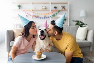 A couple wearing colorful party hats leans in to kiss their golden retriever. They are celebrating a special occasion in their cozy living room, surrounded by decorations and a treat on the table.